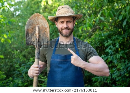 Happy farmer man in farmers hat and apron pointing finger at garden spade natural outdoors