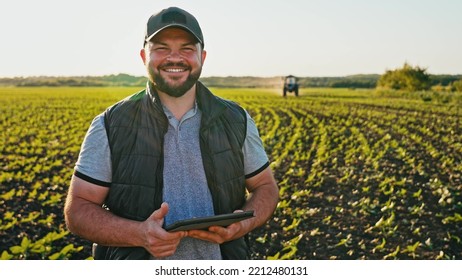 Happy farmer stands and smile holds tablet in his hands against background of working tractor in field. Concept ecology, transport, outdoor nature, clean air, food. Natural production bio product. - Shutterstock ID 2212480131