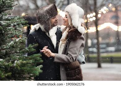 Happy Fashion Couple Of Lovers Hugging, Walking On Winter Street In Snowy Weather, Man And Woman Wear Fur Hat And Warm Wool Coat. St. Valentines Day, Christmas Holidays Concept. 