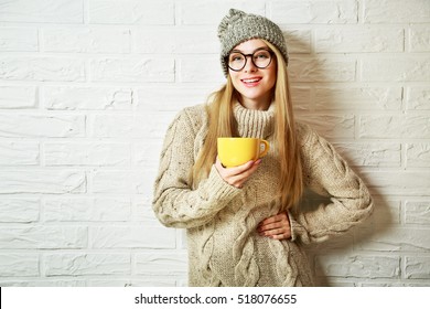 Happy Fashion Hipster Girl in Knitted Sweater and Beanie Hat with a Mug in Hands. Smiling Nice Woman at White Brick Wall Background. Winter or Autumn Warming Up Concept. Toned Photo with Copy Space.