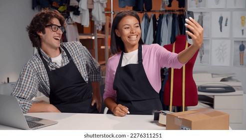 Happy fashion store warehouse workers having fun and taking selfie on smartphone. African and caucasian managers taking photo together on cellphone working in tailor storehouse