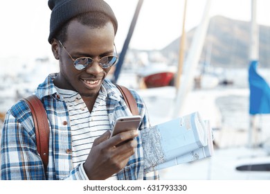 Happy fashionable dark-skinned man traveling in European resort town alone carrying paper map under his arm looking for cafe and hostels nearby using 3g or 4g internet connection on his mobile phone