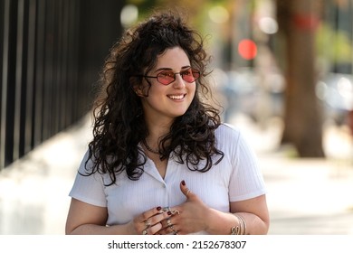 Happy fashionable woman wearing sliver jewelry and sunglasses, smiling, walking on the city street. Beautiful young girl with curly hairstyle. real people emotions. Lifestyle. Tourism.