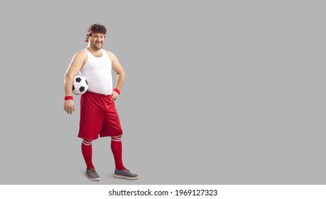 Happy Fat Man Holding Soccer Ball Under Arm. Full Body Funny Smiling Chubby Male Football Player In Headband, White Tank And Red Shorts Standing Akimbo On Free Empty Copyspace Advertising Background