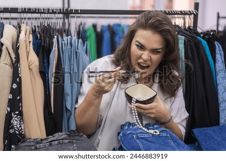 Happy fat woman fooling around and pretending to eat a pearl necklace with Chinese chopsticks in a clothing store for large people. 