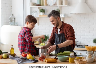 Happy Father And Adorable Son Prepare Water For Cooking Pasta. Smiling Dad Holding Pan. Kid Sitting On Table. Ingredients For Making Dinner On Table. Home Kitchen Interior. Parenthood And Childhood