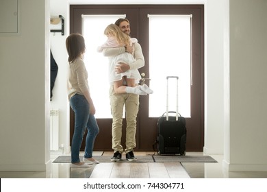 Happy father arrived home returning after business trip with baggage, daddy missed little daughter holding in arms hugging girl while wife standing in hall, family reunion, welcome back dad concept 