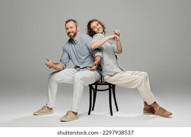 happy father and carefree teenage son having fun and pushing each other while sitting on same chair on grey