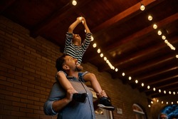Happy Father Carrying His Son On His Shoulders While Turning On The Lightbulb On A Patio.