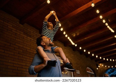 Happy father carrying his son on his shoulders while turning on the lightbulb on a patio. - Shutterstock ID 2267320123
