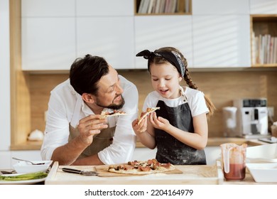 Happy father with happy child in aprons tasting eating delicious tasty homemade pizza in modern kitchen. Delighted parent looking at daughter smiling. Family spending time together having fun. - Powered by Shutterstock