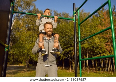 Happy father and child doing fitness exercises on sports play ground area among green trees in summer. Strong smiling dad holding son who is sitting on his shoulders and grabbing at metal chin up bar