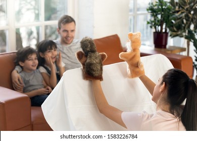 Happy Father With Cute Siblings Watching Mom Puppet Show. Smiling Young Family Sitting Together On Sofa At Home, Mother Play Muppet Toys Do Theatre Performance For Small Children.