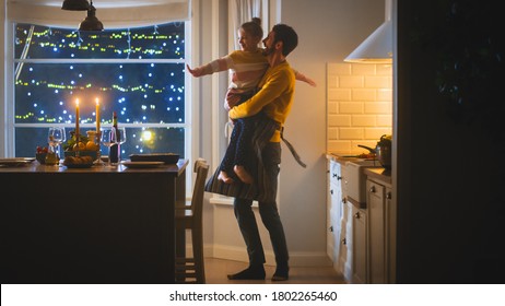 Happy Father And Daughter Cooking And Having Dinner Together. Father Prepares Food, Hugs Cute Little Girl. Festive Table In Stylish Kitchen Interior With Warm Light