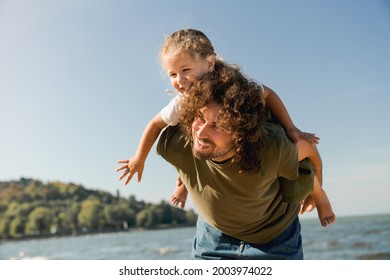 Happy father and daughter playing and having fun together on beach. Caucasian dad holding little kid child piggyback on weekend walking along lake river - Powered by Shutterstock