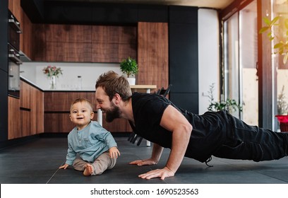 Happy Father Doing Push Ups Next To His Infant Baby At Home. Looking At His Baby, Moved And Amused By Him. Family Quarantine, Domestic Life In Self-isolation. Sunset Light From The Windows