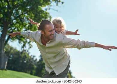 Happy Father Giving His Little Son Piggy Back Ride In Park, Pretending To Fly. Fatherhood, Childhood, Happiness Concept