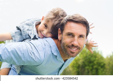 Happy Father Giving Piggyback Ride To His Son And Looking At Camera. Happy Kid Riding Piggyback On His Father. Cute Boy Flying On Father. 
