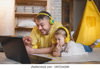 A Happy Father With His Little Daughter Is Lying On The Floor In Yellow Headphones Laughing While Looking At A Laptop. Joint Games Of Dad And Daughter