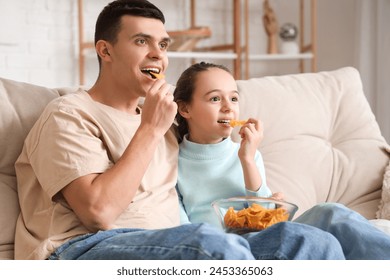 Happy father and his little daughter with nachos watching TV on sofa at home