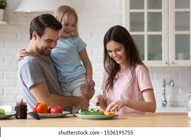 Happy Father Holding Adorable Little Daughter In Hands, Looking At Beautiful Young Mother Cooking Salad, Smiling Dad And Cute Girl Waiting For Dinner Or Breakfast, Family Enjoying Weekend Together