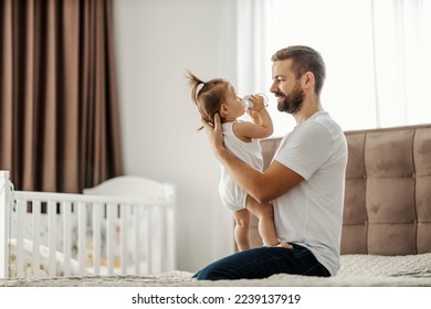 A happy father is holding his baby girl in his hands while she drinks water from a bottle. - Powered by Shutterstock