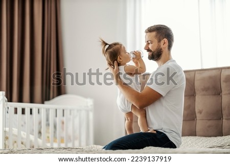 A happy father is holding his baby girl in his hands while she drinks water from a bottle.