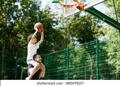 Happy father holding his little son on shoulders, helping him to score a basket on a playground