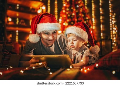 Happy father with little son in pajamas and Santa's hats lying in bed at home, uses digital tablet computer before sleeping. Christmas tree and lights in the background.  - Powered by Shutterstock