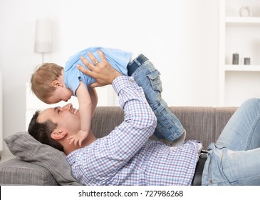 Happy Father Lying On Back On Sofa Lifting Laughing Baby Boy Above Head.