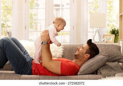 Happy Father Lying On Sofa Holding Baby Girl, Playing, Smiling. Side View.