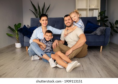 Happy father and mother with little children sitting on floor near sofa and laughing while having fun together at home during weekend 