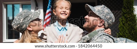 Happy father and mother in military uniforms lifting daughter while standing near house with usa flag, banner