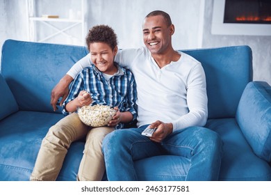 Happy father and son in casual clothes are watching TV, eating popcorn and smiling while sitting on couch at home - Powered by Shutterstock