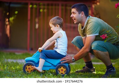 Happy Father And Son Having Fun, Playing With Bobby Car On The Home Backyard At Sunny Summer Day