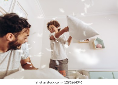 Happy Father And Son Having A Pillow Fight On Bed With Feathers Flying Around. Father And Son Having Fun Playing At Home.