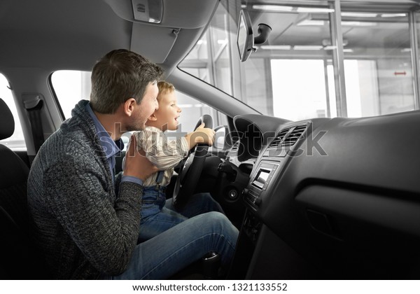 Happy\
father and son observing and testing new car in dealership. Parent\
holding kid on knees, sitting in driver\'s seat, car cabin. Little\
boy holding hands on steering wheel,\
smiling.
