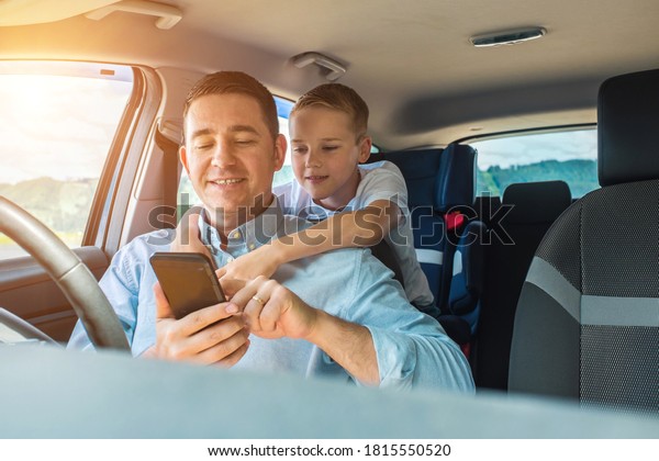 Happy father and son sitting in\
car and using smartphone in summer sunny day. Family \
travelers.