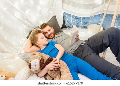 Happy father and son with teddy bear reading book in blanket fort