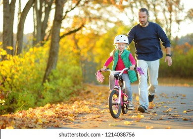 Happy Father Teaching His Little Daughter To Ride A Bicycle. Child Learning To Ride A Bike On Sunny Fall Day. Family Activities At Autumn.