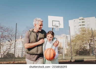 Happy father and teen daughter embracing and looking at camera outside at basketball court.