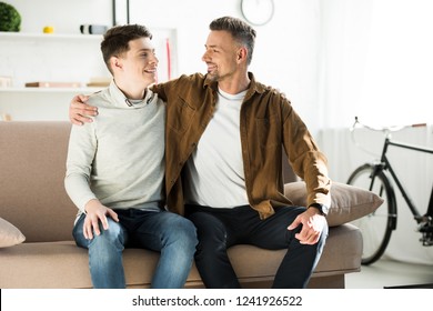 Happy Father And Teen Son Hugging On Sofa At Home, Looking At Each Other