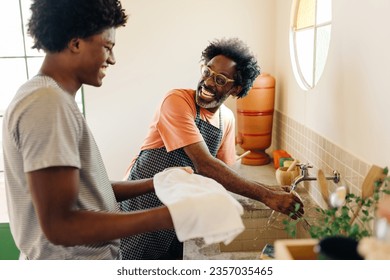 Happy father washing his hands with clean tap water while his son holds a drying towel for him. Afro-Brazilian family practicing hygiene with clay filter water in their home kitchen. - Powered by Shutterstock