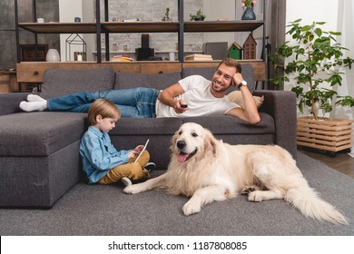 Happy Father Watching Tv On Couch While His Son Using Tablet On Floor With Golden Retriever