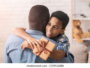 Happy Father's Day. Happy Black Boy Hugging Dad And Giving Him Gift Box At Home, Rear View Of Daddy's Back, Free Space