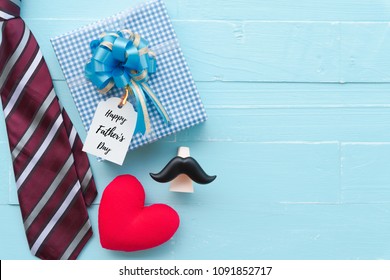 Happy Fathers Day Concept. Red Tie, Glasses, Mustache, Gift Box With Happy Father's Day Text And Handmade Red Heart On Bright Blue Pastel Wooden Table Background.