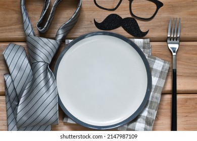 Happy Fathers day concept. Table setting - plate, napkin, tie, funny mustache with eyeglasses and cutlery on wooden table. Funny face, creative composition. Top view, flat lay, copy space.