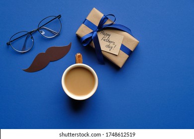 Happy Father's Day Concept. Top View Gift Box Wrapped Blue Ribbon, Greeting Card, Cup Of Coffee, Mustache And Glasses On Blue Table. Fathers Day Banner Mockup.