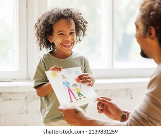 Happy Fathers Day. Cute little african american child son congratulating dad and giving him drawing with happy family on paper, smiling daddy and kid celebrating holiday at home - Powered by Shutterstock