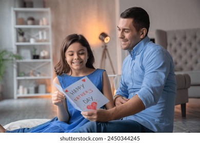 Happy Father's Day! Cute little preteen girl daughter greeting and gives handmade paper card for her attractive young father dad . Family sitting on the floor, cuddling and reading congratulations - Powered by Shutterstock
